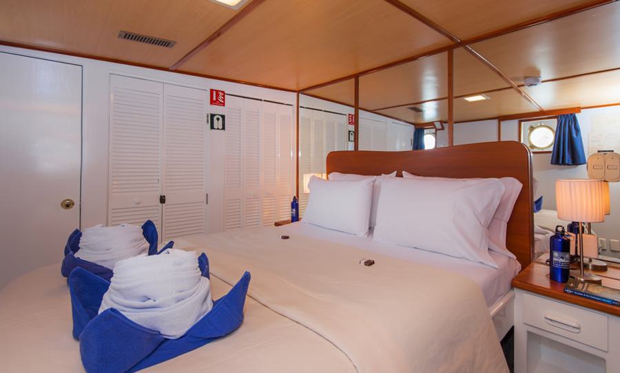 A cozy cabin with a double bed on a Beluga Galapagos cruise ship. The bed is neatly made with white linens and has two towel sculptures on it. Wooden walls and a porthole add to the nautical theme. Two bedside lamps and a couple of bottles of water are visible on the side tables.