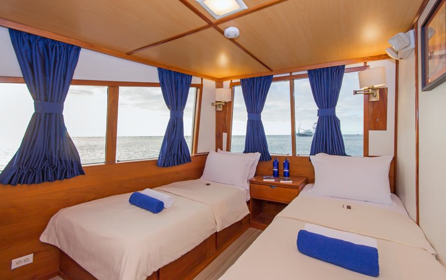 A cozy cabin on the Beluga Galapagos cruise offers two single beds covered in white linens and blue towels placed on each bed. The room features large windows with blue curtains providing a scenic ocean view. A nightstand with water bottles and a lamp is positioned between the beds.