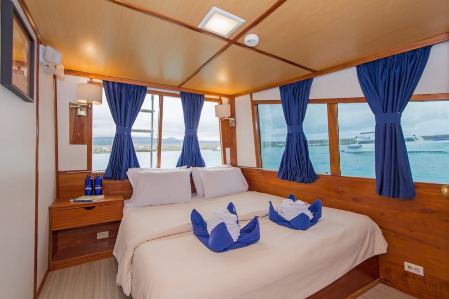 A cozy nautical-themed bedroom with a double bed, adorned with white bedding and blue towel decorations shaped like animals. The room has wooden walls and large windows with blue curtains, providing a view of the turquoise sea and a docked boat outside—perfect for your upcoming Beluga Galapagos cruise.