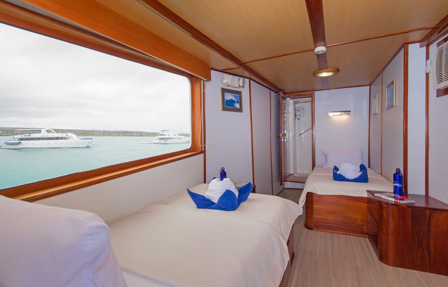 A cozy boat cabin with two single beds, each adorned with white linens and blue towels folded in a decorative manner. A window offers a view of the sea and nearby boats. The compact room on the Beluga Galapagos cruise features wood paneling, an air conditioner, a small table, and a door leading to the bathroom.