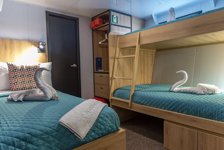 A cozy cabin room on the Bonita Galapagos cruise features a twin bed and a bunk bed with teal quilts. Towels folded into swan shapes rest on the beds. The space includes a closed black door, a small set of drawers, and an open shelving unit with orange life vests and a blue item stored inside.