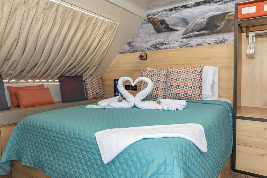 A cozy bedroom with a double bed adorned with a teal quilt, patterned pillows, and neatly folded swan-shaped towels. A mural of sea turtles from the Bonita Galapagos cruise decorates the wall above the bed. The room includes a window with beige curtains, a bench with cushions, and an open wardrobe.