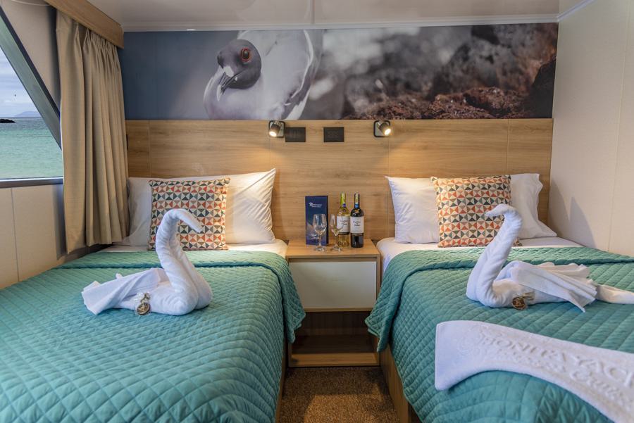 A cozy cabin with two single beds adorned with teal quilts and decorative pillows. Towels folded into swan shapes rest on each bed. Above the wooden nightstand between the beds is a wall mural featuring a bird. The window offers a sea view, perfect for guests on the Bonita Galapagos cruise.