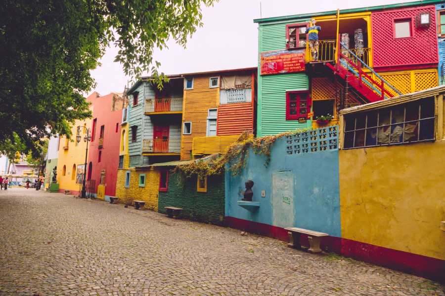 Vibrant, colorful buildings line a cobblestone street, each painted in shades of red, yellow, blue, and green. A staircase on the right leads to an upper level. Benches and greenery add charm to the scene, under a cloudy sky.