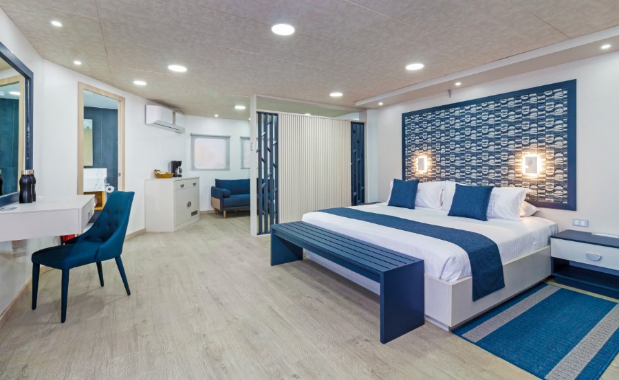 Modern hotel room with a large bed featuring white and blue bedding. The room includes a blue chair by a white desk, a seating area with a sofa, and a small kitchenette. The decor is contemporary with a blue and white color scheme and wooden flooring, perfect for guests returning from the Cormorant II Galapagos cruise.