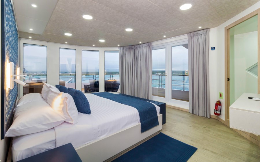 A modern hotel room features a large bed with a white and blue color scheme. The room has floor-to-ceiling windows and glass doors leading to a balcony with outdoor seating and an ocean view, perfect for resting after your Cormorant II Galapagos cruise. The interior is well-lit and decorated with minimalist furniture.