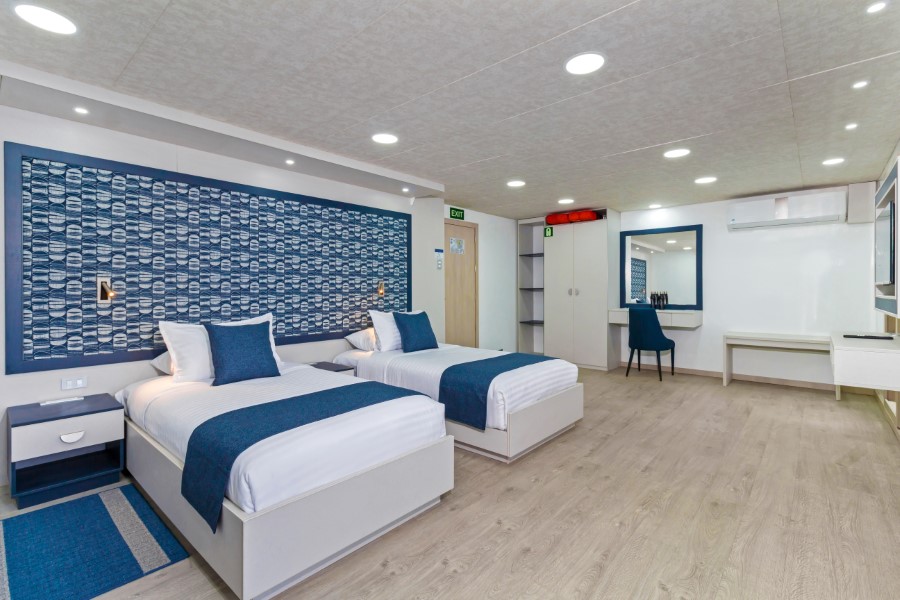 A modern hotel room with two single beds side by side, each with white linens and blue accent pillows and throws. The room features a blue patterned wall, a desk with a chair, built-in wardrobes, air conditioning, and wood flooring; perfect for guests preparing for the Cormorant II Galapagos cruise.