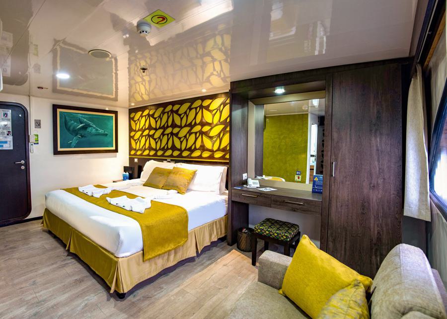 A spacious, modern bedroom in warm tones features a king-sized bed with a yellow throw blanket and pillows. There’s a decorative wall panel above the bed, a painting of a shark on the opposite wall, a dresser with a mirror, and a sofa by a large window offering views perfect for dreaming about your next Galapagos cruise.