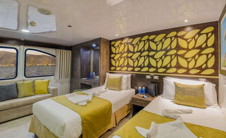 A well-lit cabin with two twin beds adorned with yellow and white bedding. The wall behind the beds features a decorative yellow leaf pattern. A sofa with grey and yellow pillows is on the left, beside large windows offering a view of the water and mountains, perfect for your Galapagos cruise on the Eco Galaxy.