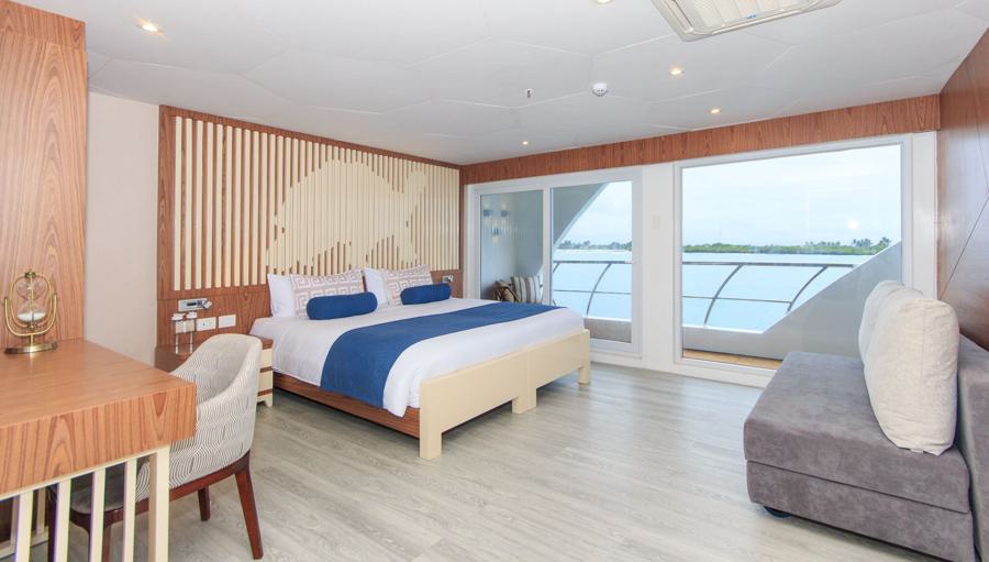 Spacious hotel room with a large bed dressed in blue and white linens, wooden furniture, a desk with a modern chair, and a gray sofa. Large windows offer a view of the sea and sky. Room features contemporary decor with wooden accents and light-hued flooring, perfect for guests before or after an Elite Galapagos cruise.