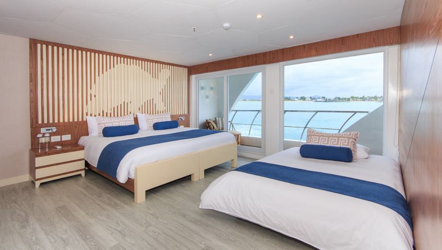 A well-lit hotel room featuring two neatly made beds with blue and white bedding. The room has a wooden floor, a bedside table, and large windows with an ocean view. The walls have a modern design with wooden accents and minimalist decor, perfect for guests starting their Elite Galapagos cruise.