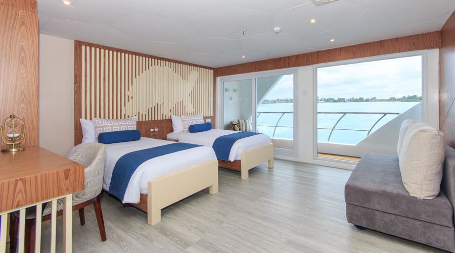 A modern hotel room features two single beds with blue throws, a desk with a chair, a couch, and large floor-to-ceiling windows offering a scenic view of a body of water. Natural light brightens the space through the expansive windows, creating an ambiance akin to an Elite Galapagos cruise.