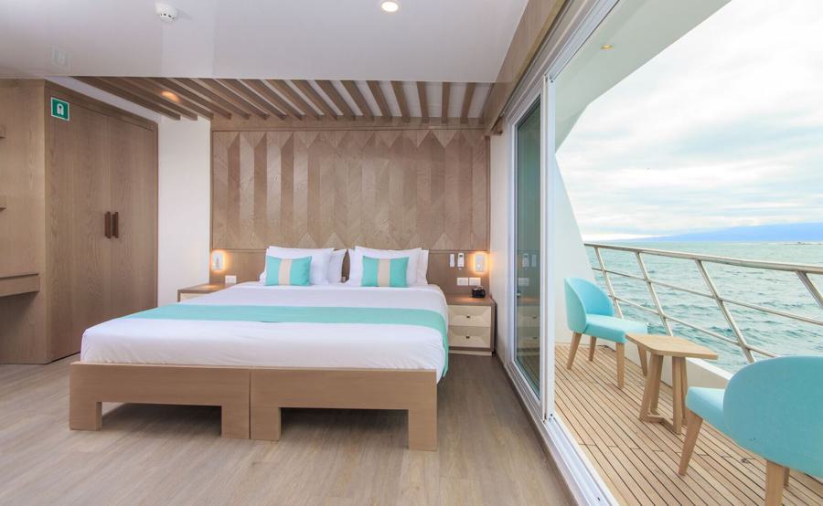 A modern, well-lit bedroom with wooden furnishings. The room features a large bed with turquoise pillows and a white duvet. A wooden wall design stands behind the bed. Sliding glass doors open to a balcony with turquoise chairs, offering a sea view, perfect for relaxing after an Endemic Galapagos cruise.