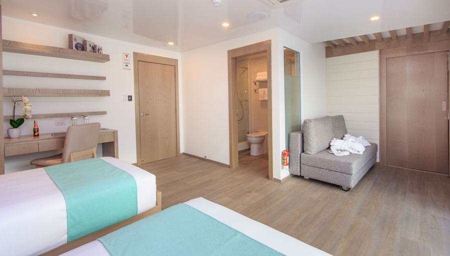 A modern hotel room with twin beds adorned with teal bedding. There's a desk and chair, decorative shelves, and a small sitting area with a couch. An open bathroom door reveals a clean, well-lit bathroom. The room features wooden floors and contemporary design elements, perfect for relaxing after an endemic Galapagos cruise.