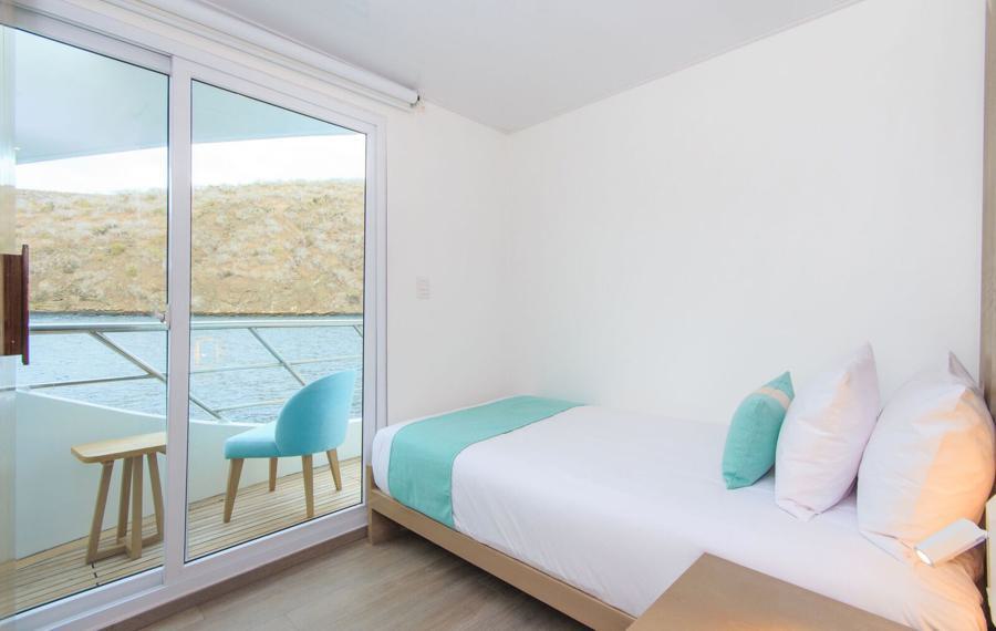 A minimalist bedroom with white walls features a single bed adorned with white and turquoise bedding. A glass sliding door opens to a balcony overlooking a water body, furnishing a light blue chair, a wooden stool, and a scenic rock wall in the background. The decor echoes the tranquility of an Endemic Galapagos cruise.