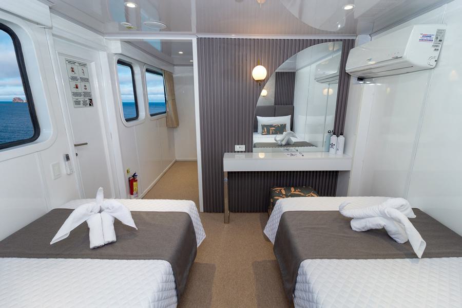 A clean, modern cabin room on a ship with two single beds dressed in white linens and gray throws. Each bed has a towel folded in the shape of a bird. The room has a large round mirror, an air conditioner, and windows with ocean views.