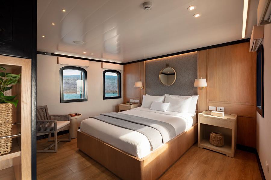 A cozy, modern bedroom on a Grace Galapagos cruise boat features wooden decor. A large bed with white linens is centered against a wall, flanked by two nightstands with lamps. A round mirror hangs above the bed, and two windows offer views of the water. A chair and small table sit in one corner.