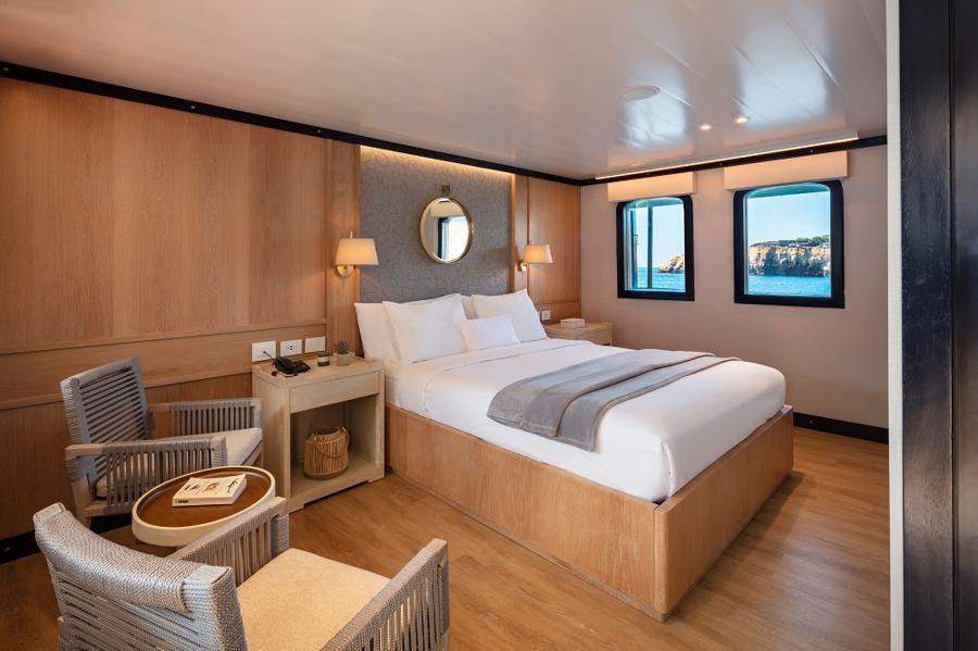 Bright and cozy cabin interior on the Grace Galapagos cruise with a large window view of the sea. Features a neatly made double bed, wooden furniture, a small round table, two chairs, wall lamps, a round mirror above the bed's headboard, and a woven basket under the desk.