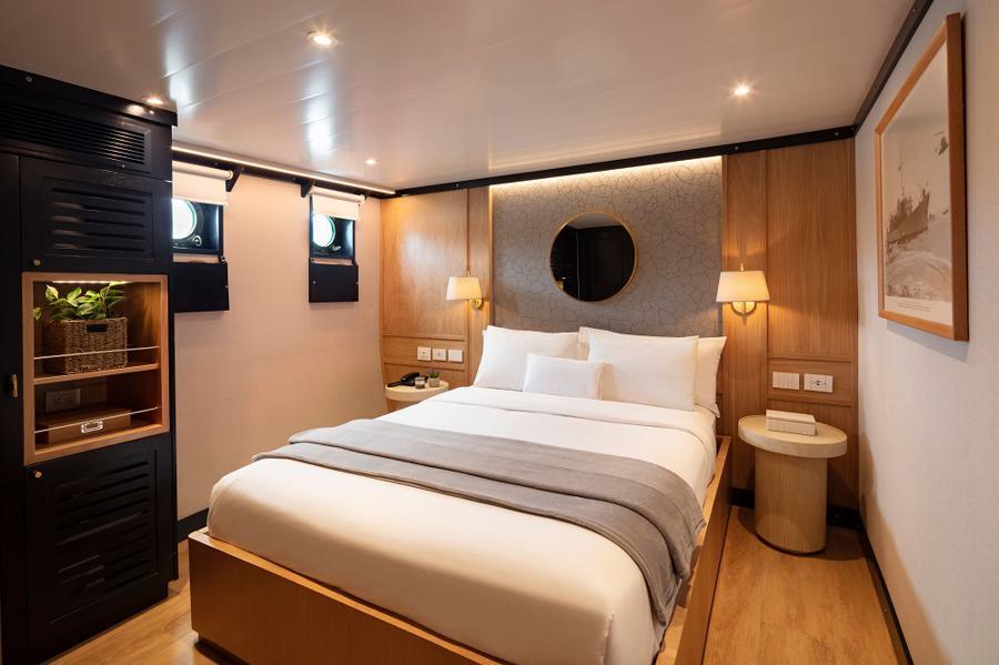 A cozy, well-lit ship cabin on the Grace Galapagos cruise features a double bed neatly made with white linens and a gray throw. The wooden walls have a round mirror and a framed picture. Two small round windows, a side table, and a closet with plants add charm to the space.