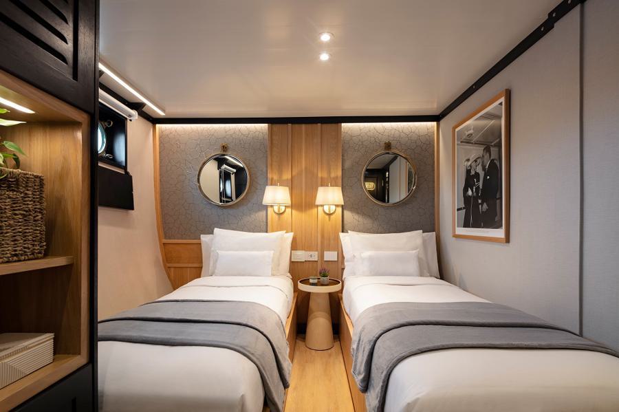 A cozy, modern hotel room with twin beds dressed in white linens and gray blankets. The walls are adorned with round mirrors and a black-and-white portrait. Between the beds is a small wooden nightstand with a phone and two wall-mounted lamps. On the left, there is a wooden storage unit with wicker baskets and decor, much like you'd find on a Grace Galapagos cruise.