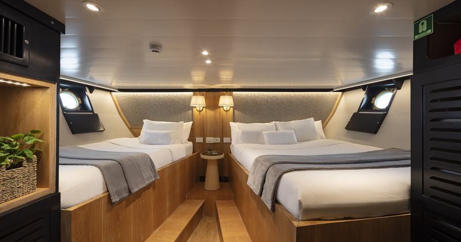 A cozy cabin room with two neatly made beds, each with white linens and gray blankets. The beds are arranged at an angle, with a nightstand and two lamps between them. The room features wooden accents, a small plant, circular windows, and soft overhead lighting—perfect for your Grace Galapagos cruise.