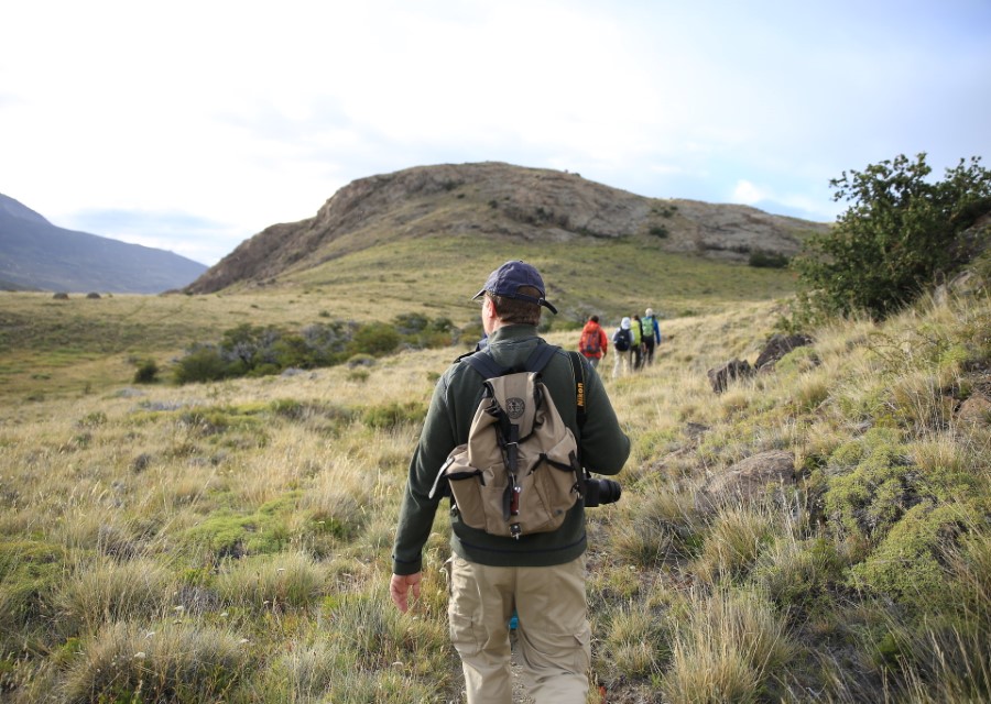 A group of hikers walks through grassy hills on a clear day. The closest hiker, wearing a cap, a backpack, and a green jacket, leads the way. Rolling hills and sparse vegetation expand into the distance.