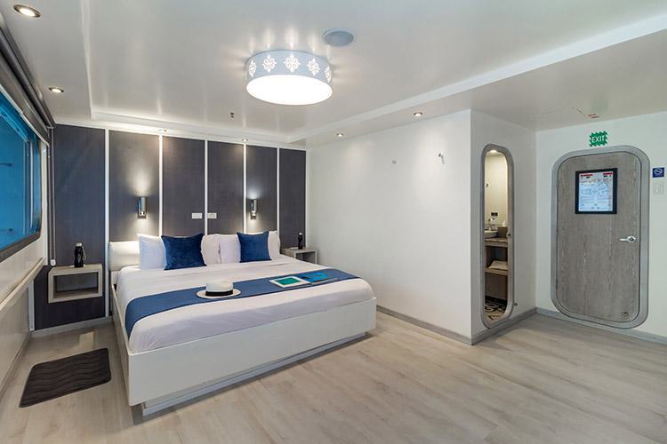 A modern hotel room with a large bed, decorated in white and gray tones. The bed has white linens and blue accent pillows, with a hat and tablet on it. A circular ceiling light fixture illuminates the room. An archway leads to a bathroom, and there is a door next to it—a serene retreat perfect before embarking on your Horizon Galapagos Cruise.