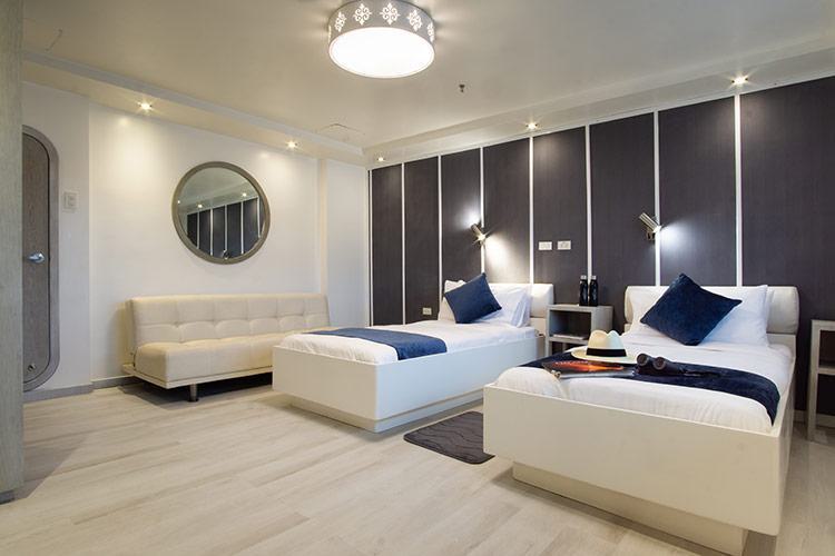 A modern hotel room with two single beds, each with white linens and navy blue accent pillows. A white sofa is placed against the left wall under a round mirror. The room features grey and white walls, wood flooring, and a stylish ceiling light fixture—perfect for guests of the Horizon Galapagos Cruise.