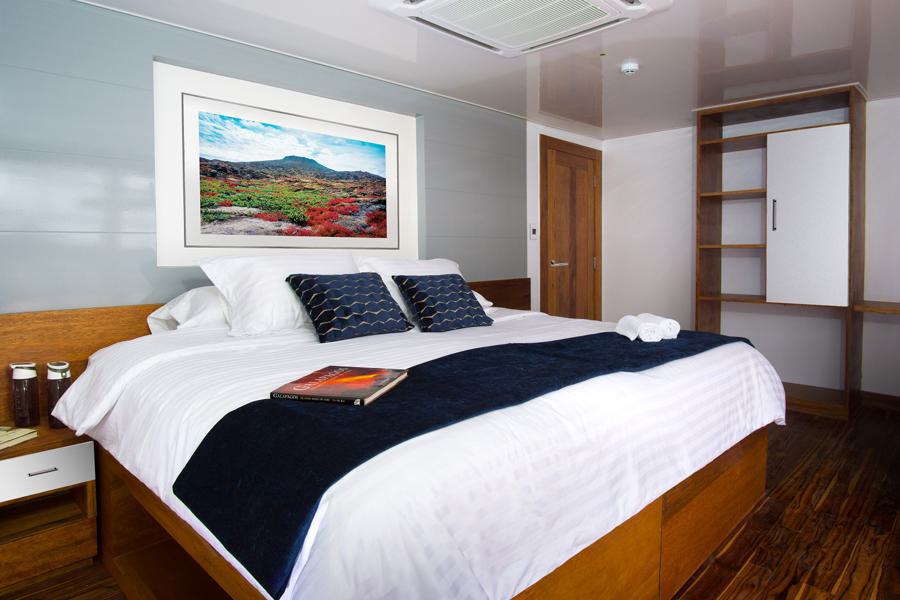 A cozy bedroom features a neatly made bed with white linens, dark blue pillows, and a matching throw. A landscape photo adorns the wall above the headboard. A wooden door, built-in shelves, and a nightstand with jars complement the modern, minimalistic decor inspired by an Infinity Galapagos cruise.