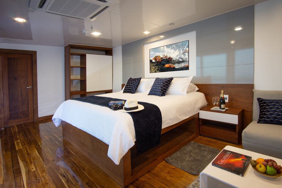 A modern, tastefully decorated bedroom with a large wooden bed featuring white and navy bedding. A white hat rests on the bed. The room has polished wooden floors, a cozy armchair, a side table with a book and fruit, and a painting above the bed—a perfect retreat after an exciting day on an Infinity Galapagos cruise.