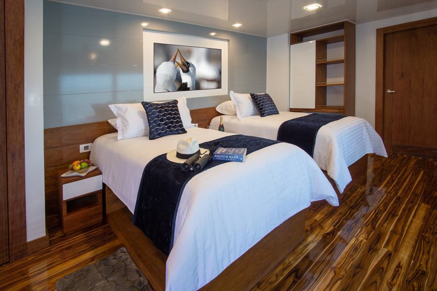 A modern bedroom with wooden floors featuring two twin beds with white linens, navy blue throws, and navy blue pillows. A hat, a book, and binoculars rest on one bed—perfect for dreaming about an Infinity Galapagos cruise. A framed picture of a horse and a small nightstand with fruits are also visible.