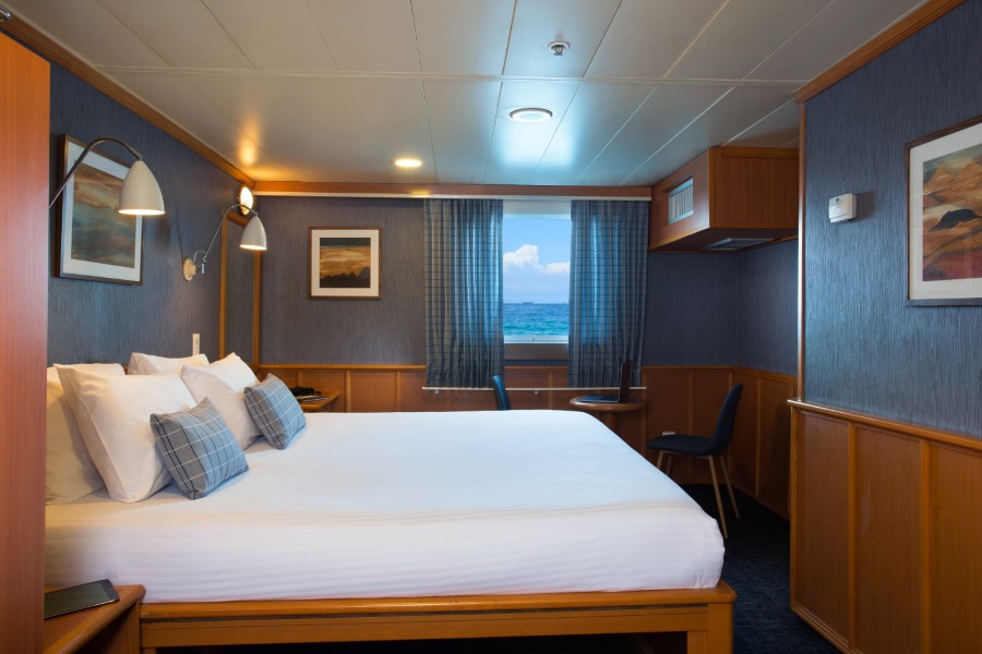 A cozy cruise ship cabin on the Isabela II Galapagos cruise features a neatly made double bed with white linens and checkered pillows. The room has wooden furniture, framed artwork, wall lamps, a small desk and chair, and a large window showcasing an ocean view.