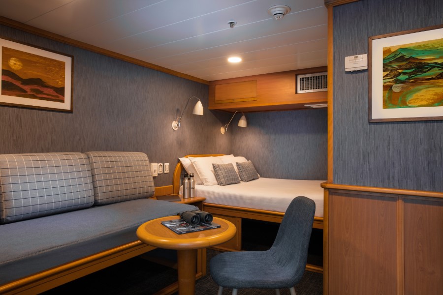 Cozy cabin room with a double bed, wall-mounted reading lights, and a small seating area with a cushioned bench and round wooden table. The room, similar to accommodations on an Isabela II Galapagos cruise, is decorated with framed artwork and has wooden paneling with a modern design.