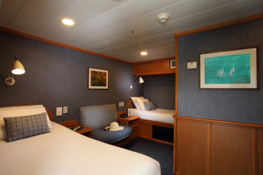 A cozy cabin room with two single beds, each neatly made with white linens and checkered pillows. The room has wood paneling, a wall-mounted lamp above each bed, a painting of sailboats from the Isabela II Galapagos cruise, and a small seating area. The overall ambiance is warm and inviting.