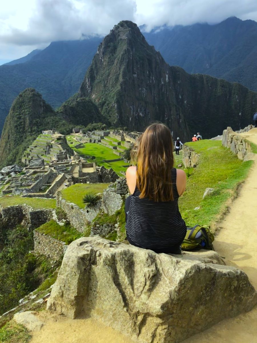 A person with long hair sits on a rock, facing the ancient ruins of Machu Picchu in Peru. Lush green terraces and stone structures are visible with a backdrop of towering mountains under a partly cloudy sky. A backpack is placed beside the person.