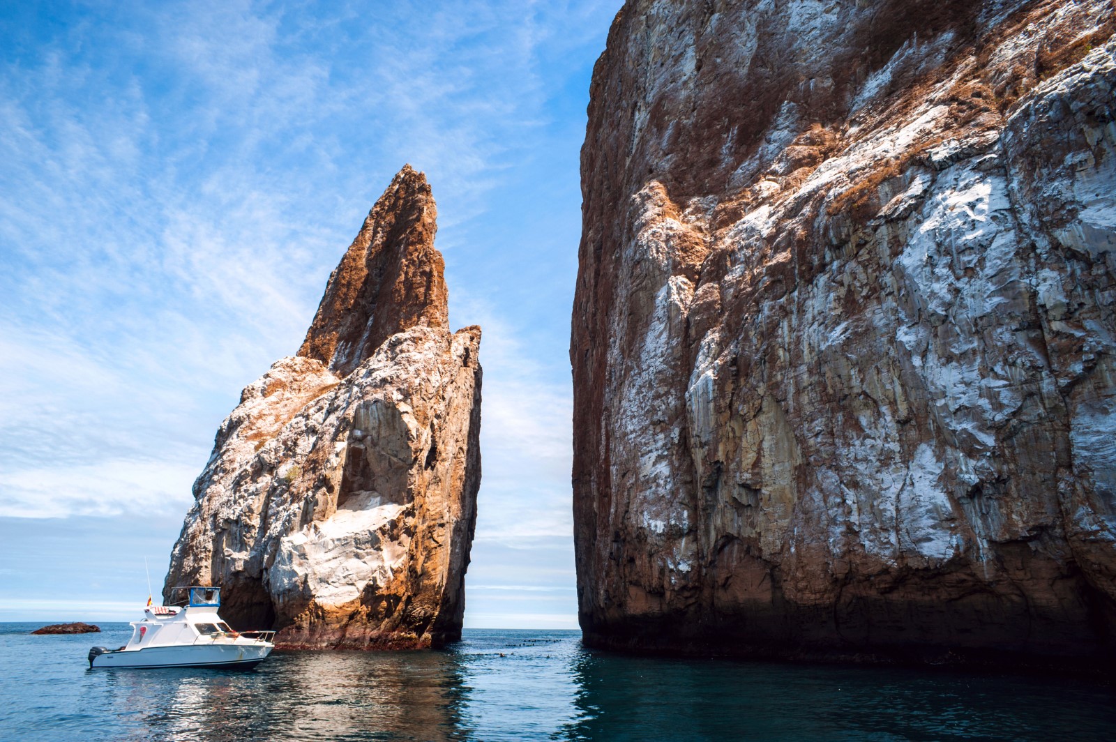 A small white boat floats on calm, blue waters near the towering Kicker Rock formation in the Galápagos Islands. The rock has steep, rugged cliffs with patches of lighter-colored stone against a backdrop of a partly cloudy sky.