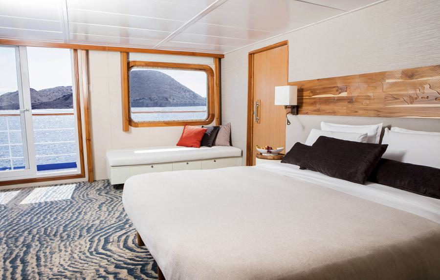 A well-lit cruise ship cabin with a large bed, a cushioned bench with pillows by a window overlooking the ocean, and a wooden door. The Legend Galapagos cruise cabin features modern decor with a patterned carpet and wooden accents. A mountainous landscape is visible through the window.