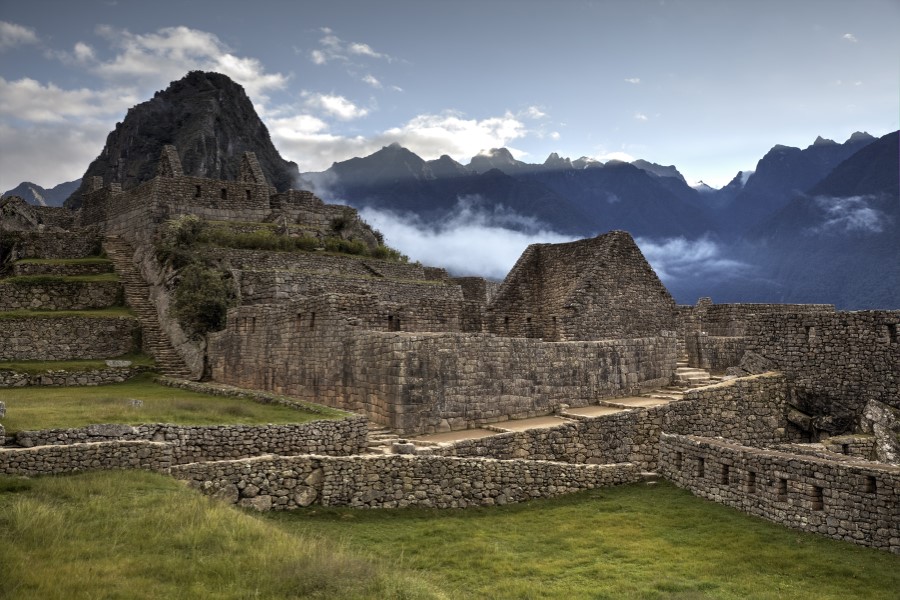 An ancient stone structure, part of the ruins at Machu Picchu, stands against a backdrop of misty mountains and a partly cloudy sky. The terraced levels and stone walls of the site are clearly visible, surrounded by lush green vegetation.