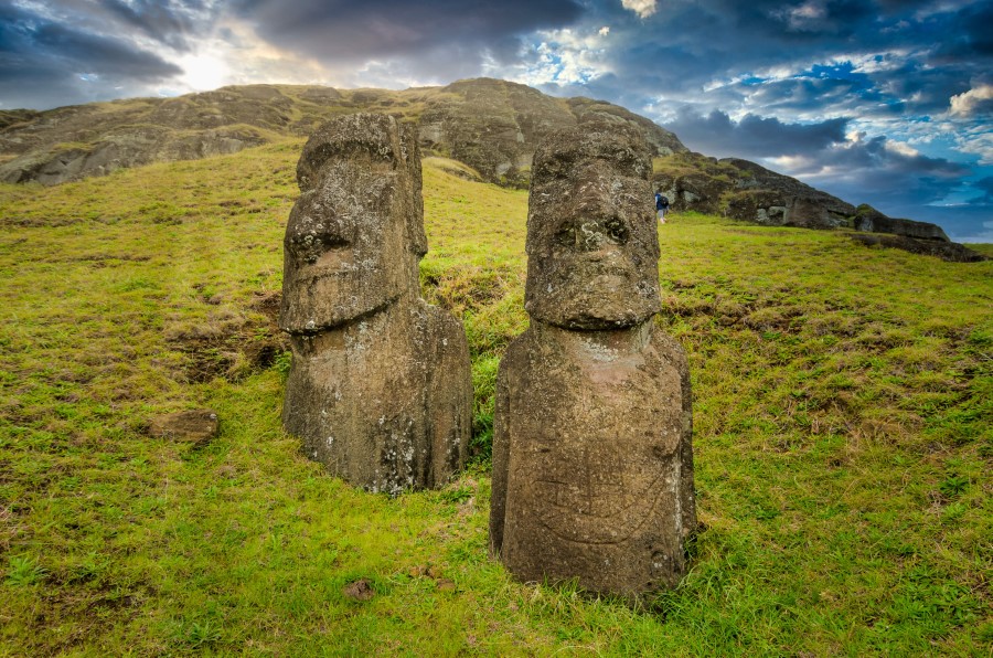 Two large stone Moai statues stand on grassy terrain with a hill and partly cloudy sky in the background. The statues, part of Easter Island's famous collection, appear weathered and ancient, embodying the island's rich cultural heritage.