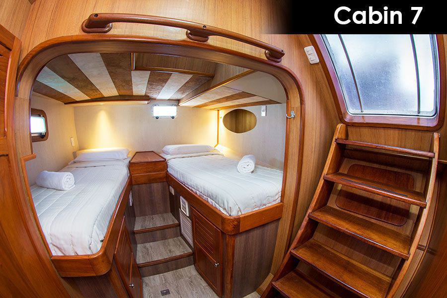 A cozy wooden cabin labeled "Cabin 7" on the Nemo Galapagos cruise features two twin beds, each with white linens and a rolled-up towel on top. The cabin has a small window, a built-in storage area, and wooden steps leading up to a frosted glass hatch. Warm lighting enhances the snug atmosphere.