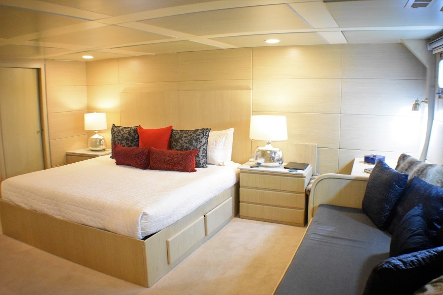 A modern bedroom features a large bed with white bedding and multiple red and grey pillows. Two bedside tables with lamps flank the bed. A cozy sofa with dark blue cushions sits beside a window on the right, offering a tranquil view that evokes the calm of a Passion Galapagos cruise. The walls and ceiling are covered with light wood panels.