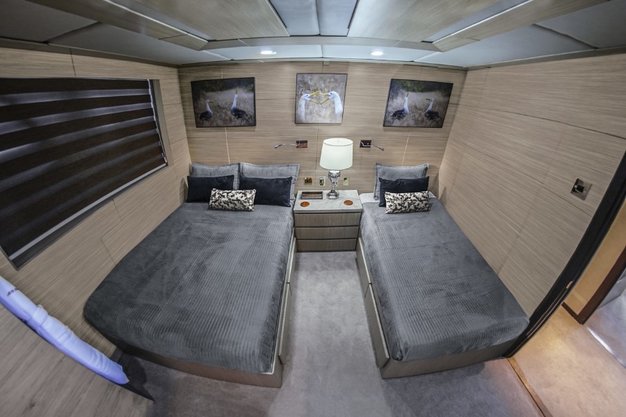 A modern bedroom with two single beds separated by a small nightstand with a lamp. The beds are dressed in dark gray linens and matching pillows. Framed artwork hangs above each bed, evoking the tranquility of a Passion Galapagos cruise. A window with horizontal blinds is on the left wall.