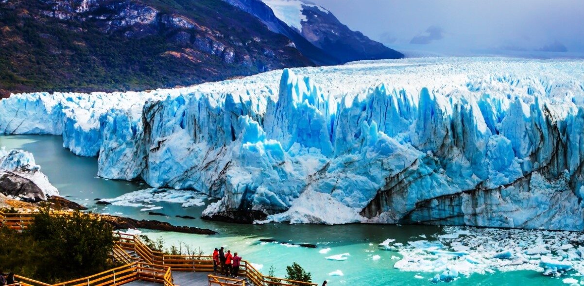 The Perito Moreno Glacier, with its jagged blue ice, towers above a milky turquoise lake in Argentina, surrounded by green mountains. Wooden boardwalks with railings provide viewpoints for visitors who admire the stunning natural scenery in the foreground.