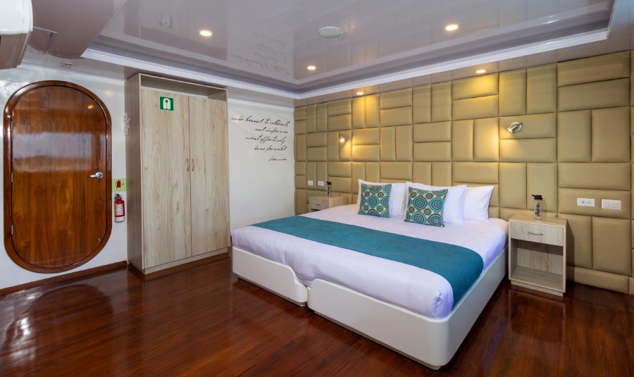 A modern hotel room with wooden flooring, a large bed with white and teal bedding, and a cushioned headboard. The room features built-in wardrobes, a round wooden door, and bedside tables with lamps. The wall above the bed has decorative writing.