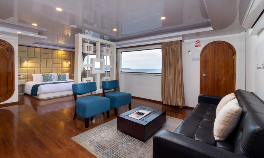 The image shows a luxurious, modern Petrel Galapagos cruise ship cabin with a sitting area and a bedroom. The space features wooden floors, a sofa, two blue chairs, a small table, and a bed with plush pillows. Large windows offer an ocean view, and the room is well-lit.