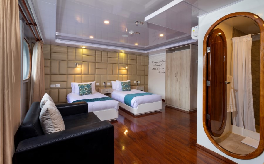 A modern, well-lit bedroom features two neatly made single beds with teal accents, a black sofa, and wooden flooring. The room includes a small wooden-framed bathroom with a shower. Large windows with beige curtains bring in natural light, ideal for relaxing after an adventurous day on a Petrel Galapagos cruise.