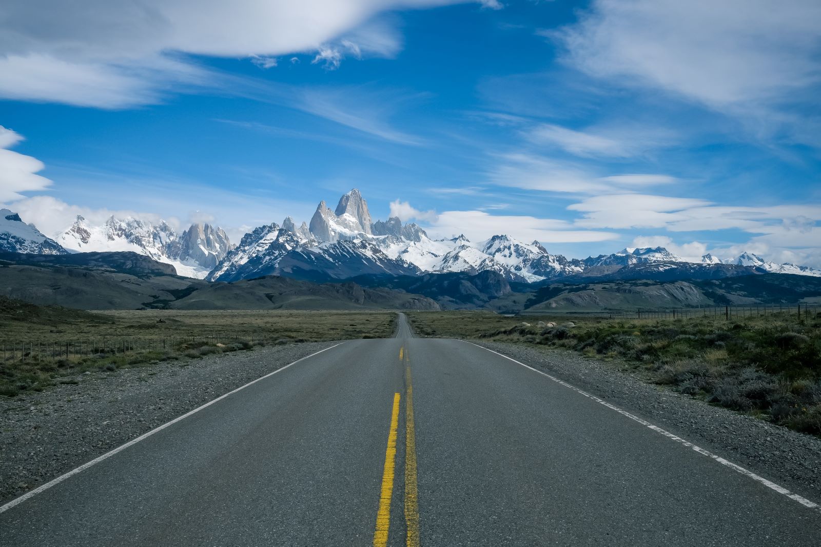 A straight road stretches into the distance towards a rugged mountain range with snow-capped peaks under a vibrant blue sky with scattered clouds. The surrounding landscape is barren, with sparse vegetation on either side of the road.