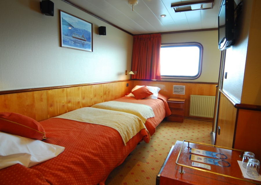 A cozy, well-lit cabin with a single bed and orange bedding, featuring a small window with red curtains, a wall-mounted picture, and a TV. A nightstand and a table with amenities are also present. The cabin has wooden paneling and beige walls, perfect for your Skorpios III Glacier Cruise in Chile.