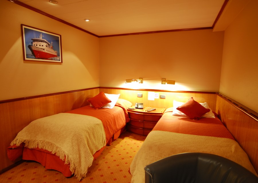 A cozy cabin room with two single beds, each adorned with orange bedspreads and beige throws. A nightstand with a reading lamp is situated between the beds. A framed picture of a ship adorns the wall above one bed, hinting at adventures like those on a Skorpios III Glacier Cruise in Chile. The room is warmly lit and comfortably furnished.