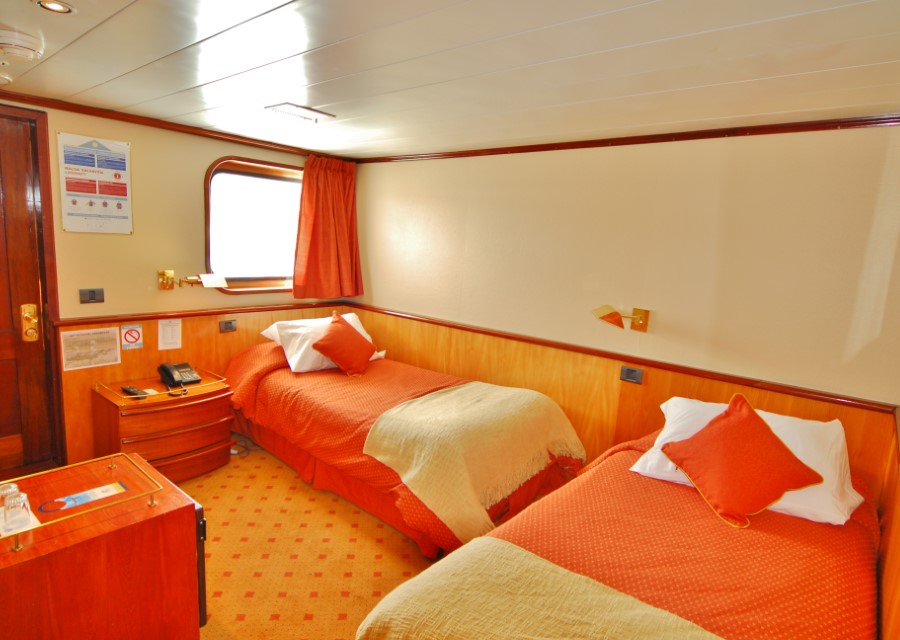 A well-lit cabin with twin beds, each adorned with orange bedspreads and a beige blanket. Matching orange pillows add color. A window with orange curtains, wooden furnishings, and a small round bedside table complete the cozy room layout—a perfect retreat on your Skorpios III Glacier Cruise.